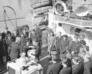 Christening of Lieutenant and Mrs. Shaw's child aboard H.M.C.S. QUINTE, at Digby, Nova Scotia, Canada on 19 September 1944. A storage locker draped with flags serves as an alter, QUINTE’s upturned bell is used as the Christening basin and shell casings from her guns are flower vases. PA-189244